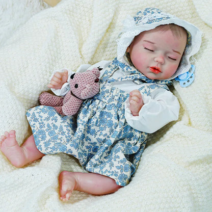 Reborn baby doll in a blue floral dress, lying on a cream blanket with one arm raised, holding a pink toy.