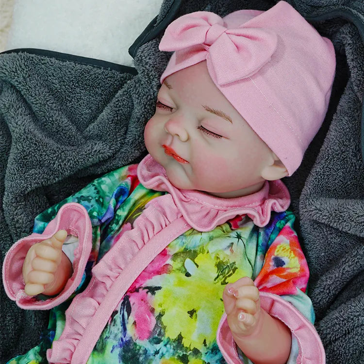 A reborn baby doll peacefully sleeping, dressed in a floral outfit with a pink headband, on a dark blanket.