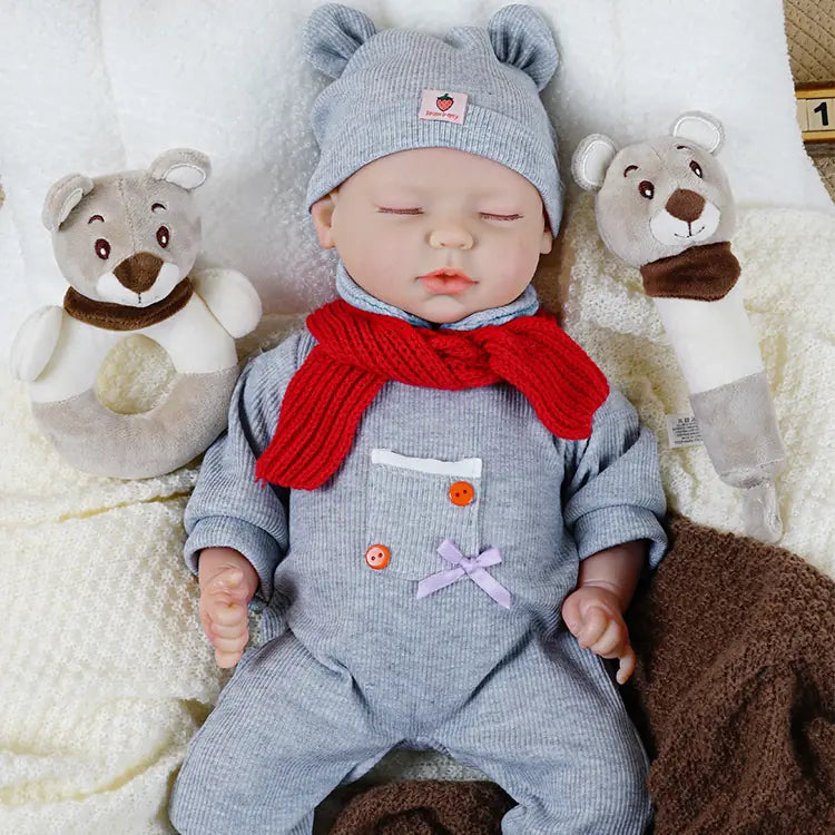 A baby doll dressed in a grey outfit with a matching hat and a red scarf. The doll is wrapped in a soft blanket, with its eyes closed and a peaceful expression, giving it a serene and lifelike appearance.