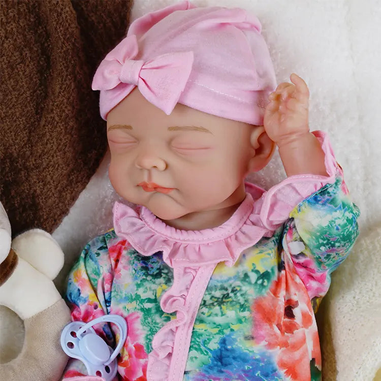 The first reborn baby doll is in a different pose, lying on her side with the cream blanket pulled up to her waist, the teddy bear, and pacifier close by.