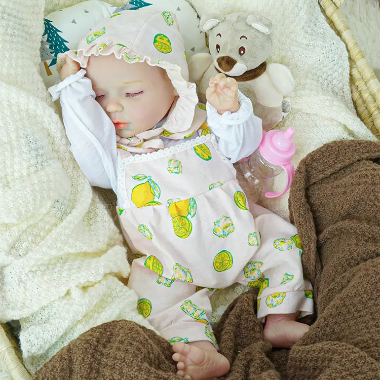 Close-up of a sleeping reborn baby doll wearing a pink bonnet and lemon-print romper. The doll rests in a woven basket with its hand gently raised, surrounded by a white fluffy blanket and a cuddly plush teddy bear.