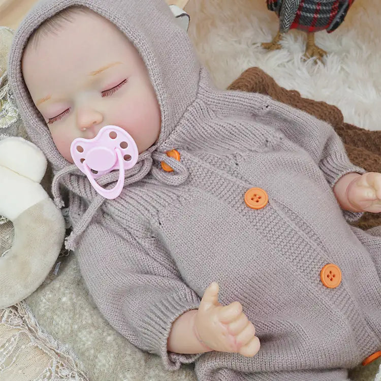 The reborn doll toy dressed in a lilac knitted jumpsuit and hood, peacefully asleep among cozy blankets and plush toys, with a teepee tent in the background.