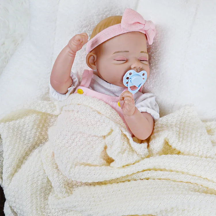 A reborn baby doll with a pink headband and blonde hair, holding a blue pacifier, and lying on a fluffy white blanket.