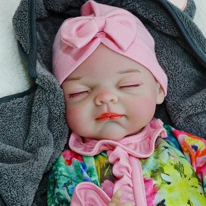 A reborn baby doll with eyes closed, dressed in a colorful floral outfit and a pink headband, resting on a dark blanket.