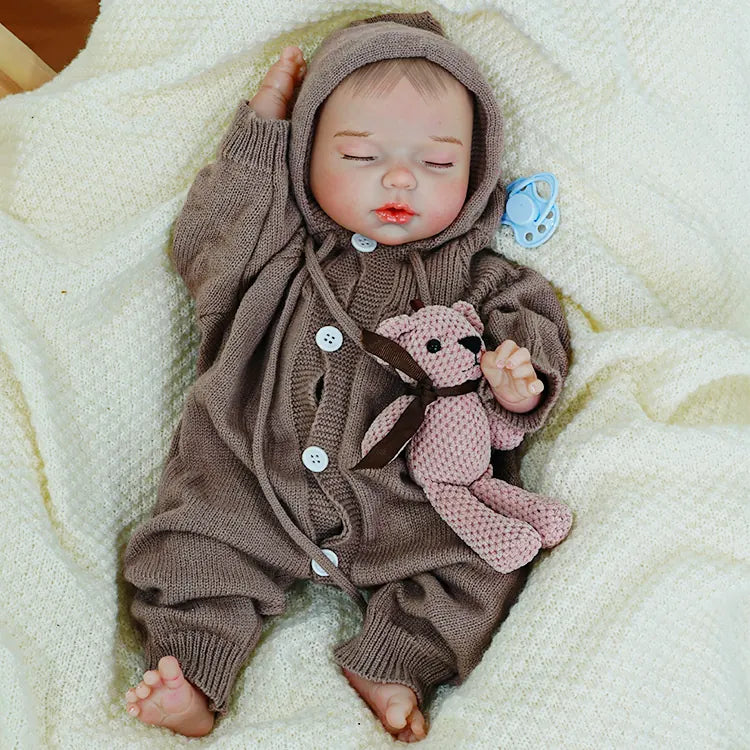 A reborn baby doll sitting upright, holding a teddy bear, quietly gazing forward.