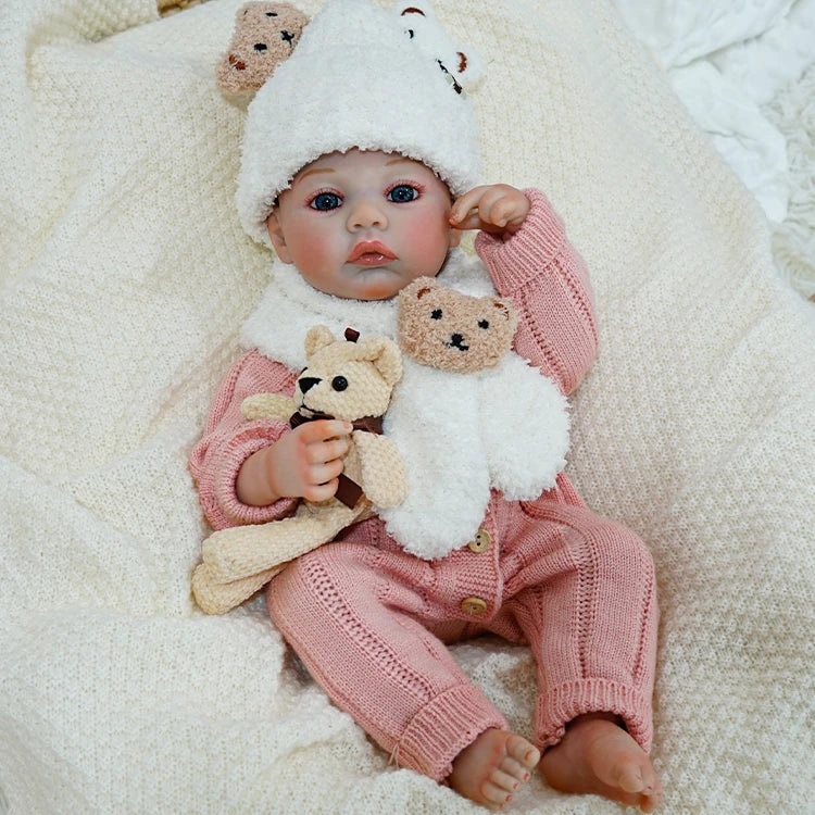 Reborn baby doll sitting, wearing a bear hat and holding a soft teddy bear.