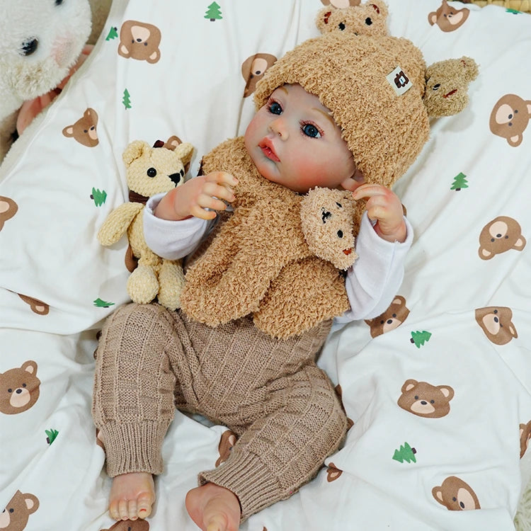 Reborn baby doll sitting with a teddy bear on a patterned blanket.