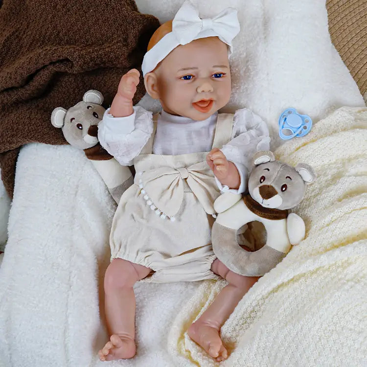 Lifelike reborn baby doll wearing a pink bow headband and a white scarf, cuddled up with a plush bear and smiling at the camera.
