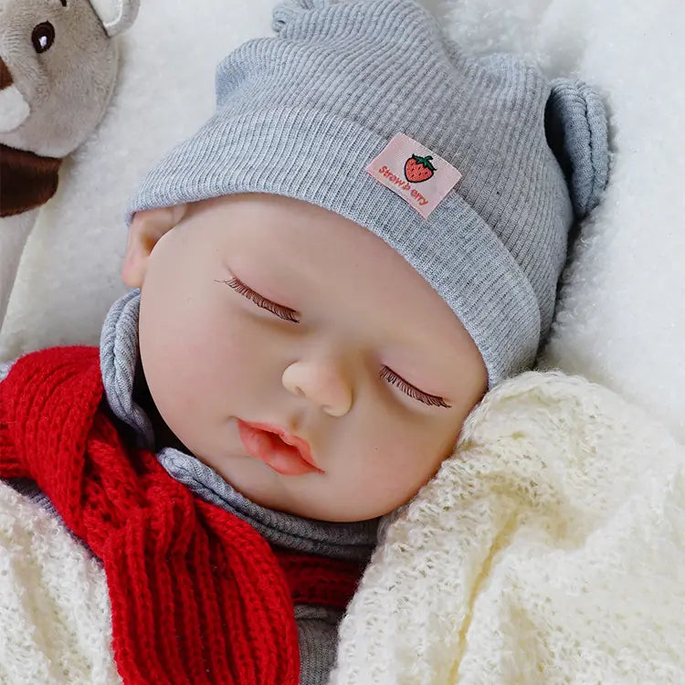 A baby doll dressed in a grey outfit with a matching hat and a red scarf. The doll has a blue pacifier in its mouth and is lying on a soft blanket with its eyes closed, creating a serene and lifelike appearance.