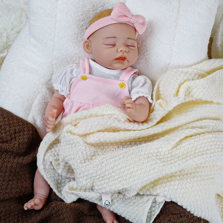 Lifelike baby doll wearing a pink and white outfit with a matching headband, peacefully sleeping on a soft, white surface.