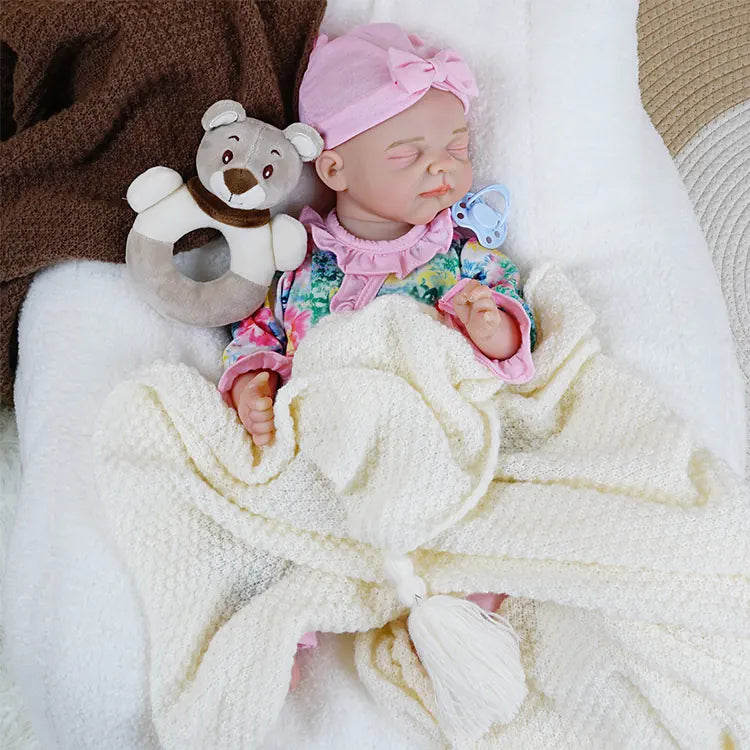 A similar pose to the previous, with the reborn baby doll lying on a fluffy white cushion, the teddy bear and blue pacifier positioned at her side.