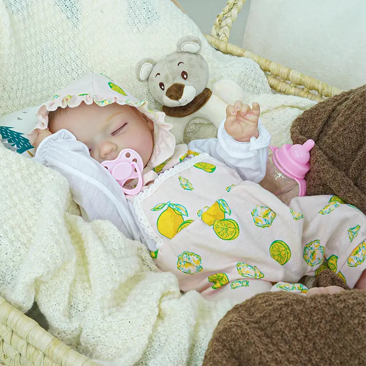 A reborn baby doll dressed in a pink lemon-print romper and matching bonnet, sleeping peacefully in a basket. Beside the doll are a plush teddy bear and a pink baby bottle, all nestled within soft, knitted blankets.