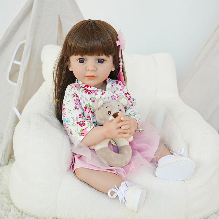 Close-up of toddler reborn doll with brown hair, wearing a floral top and holding a teddy bear, sitting on a white fluffy chair.
