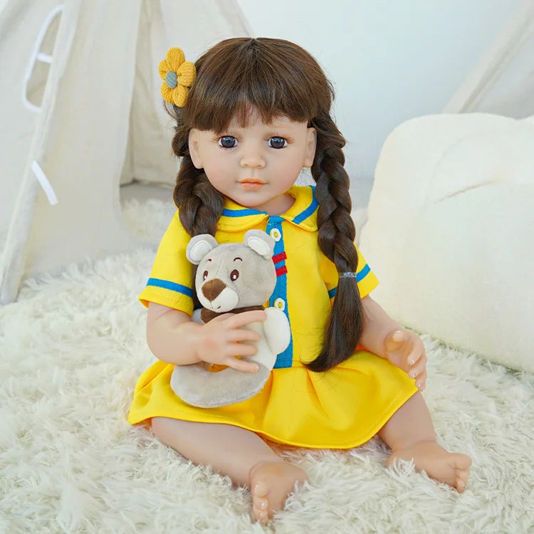 A brunette reborn baby doll with braids, wearing a yellow dress, sitting on a carpet and holding a teddy bear.