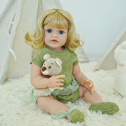 Reborn doll with blonde hair in a green dress, sitting and holding a teddy bear.