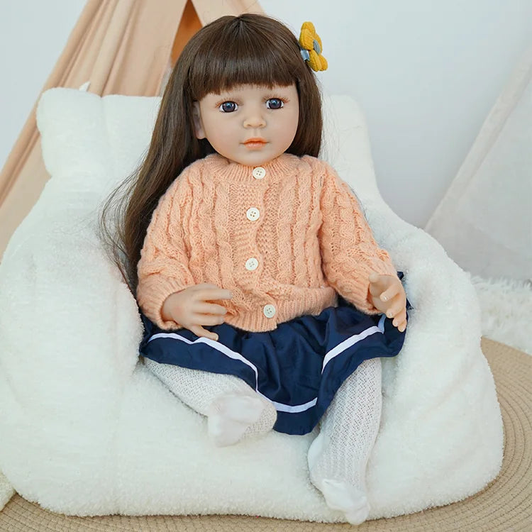 Reborn baby doll in peach sweater and navy skirt, sitting in a white chair.