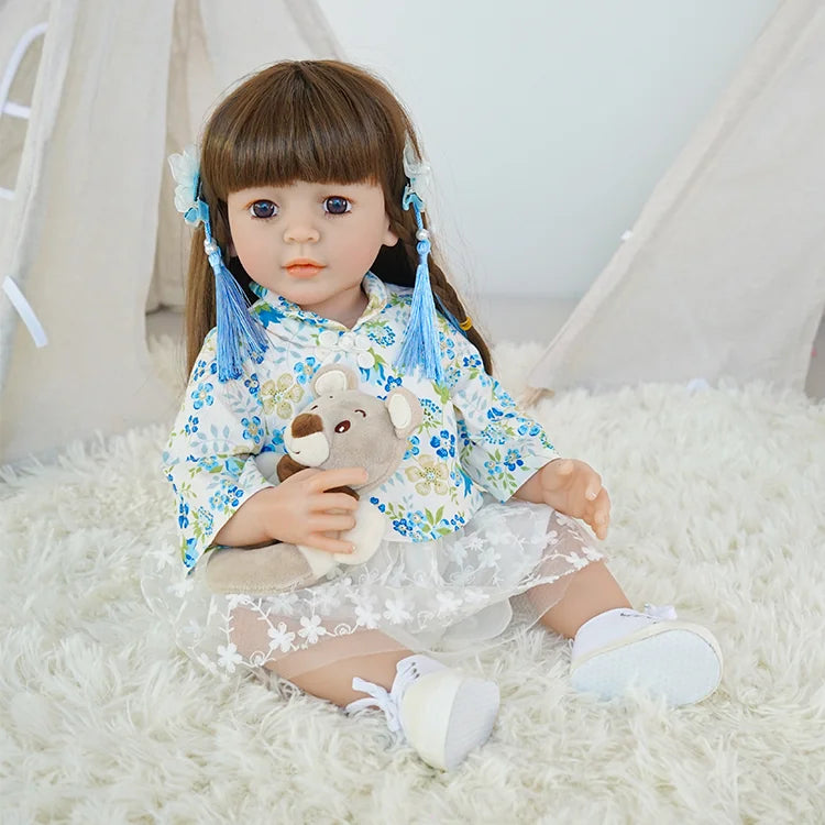 Toddler reborn doll with brown hair, dressed in a blue floral top, seated on a white fluffy chair.