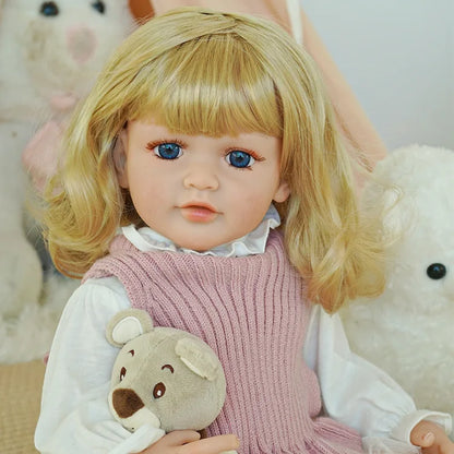 Reborn baby doll wearing a pink knit dress and white hat, holding a teddy bear.