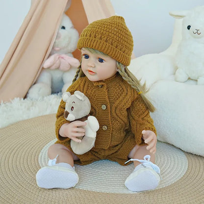 Reborn baby doll in brown knit outfit with white sneakers, holding a teddy bear.