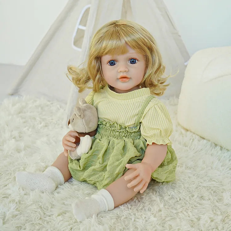 A photo of an adorable blonde curly-haired reborn baby doll wearing a light yellow top and green dress, sitting against a soft background, with bright eyes and slightly open mouth.