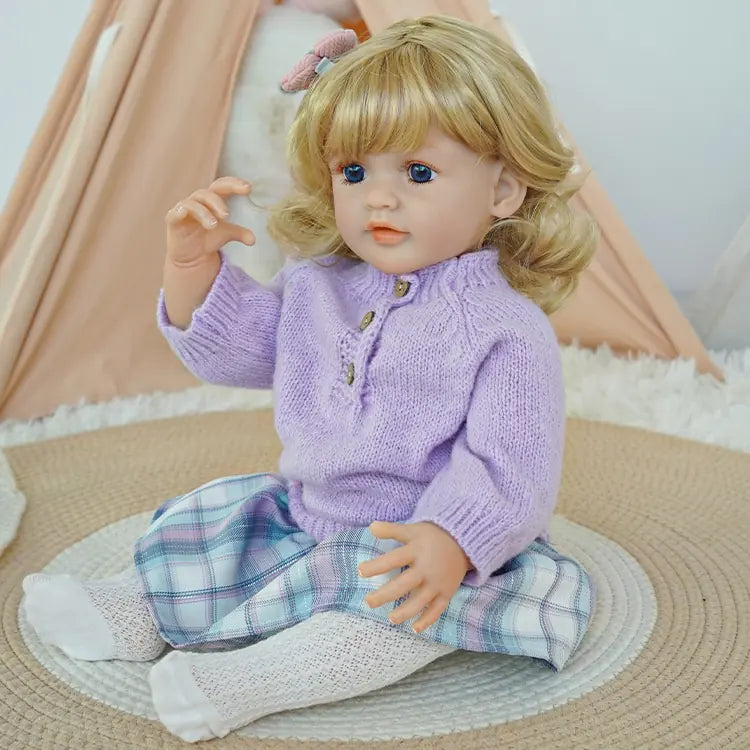 Blonde hair, blue-eyed reborn doll wearing a purple sweater and plaid skirt, holding a teddy bear, sitting on a cushion.