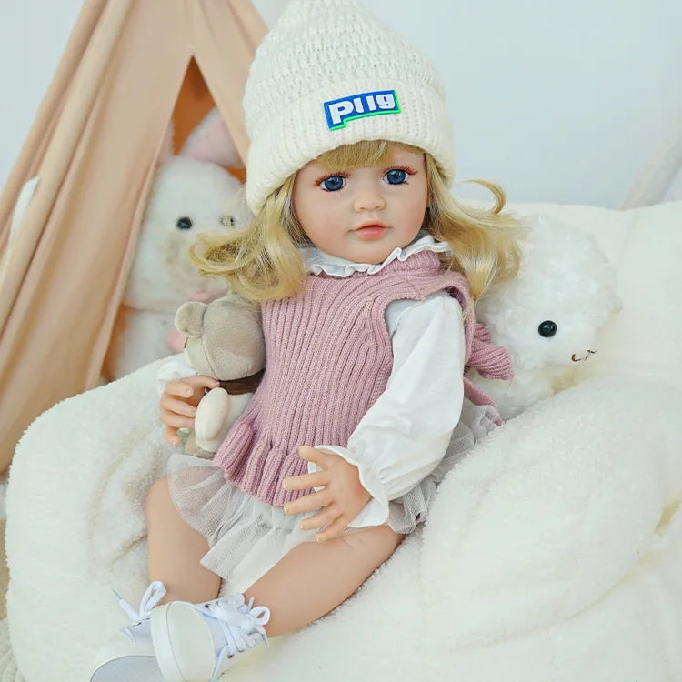 Reborn baby doll sitting in a white chair, wearing a pink knit dress and white hat.