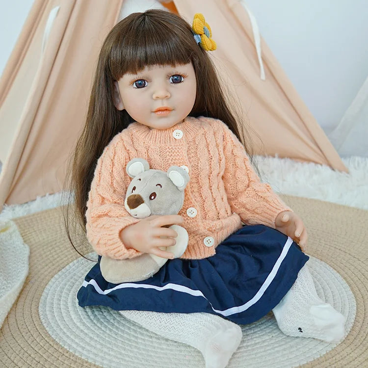 Reborn baby doll in peach sweater and navy skirt, holding a teddy bear, sitting on a rug.