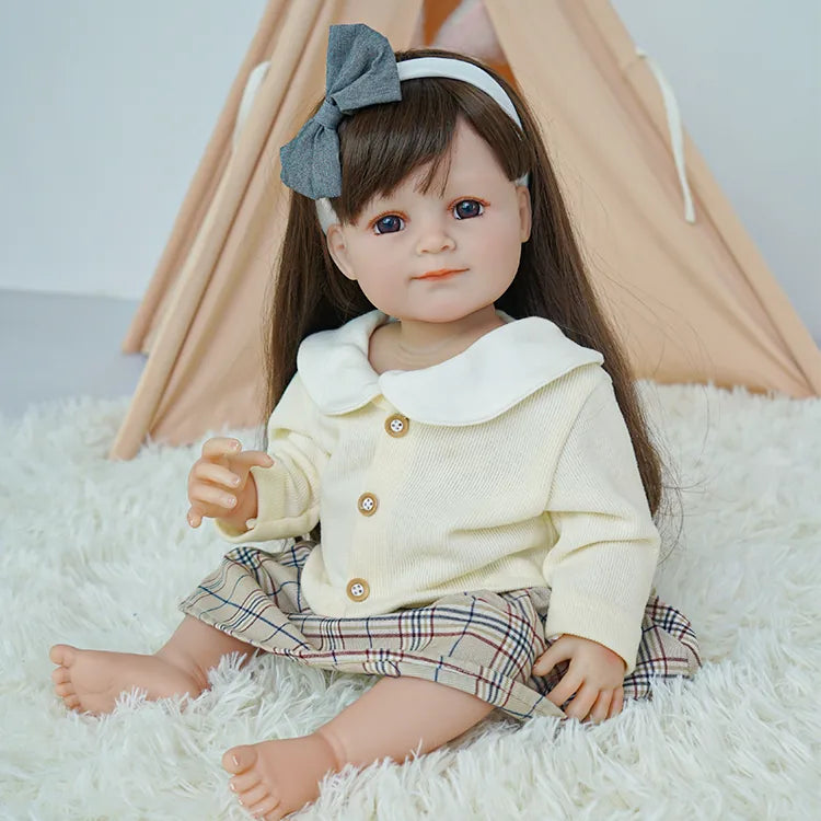 This adorable toddler doll, with her soft brown hair and sweet smile, is dressed in a cozy cream sweater and plaid skirt. The grey bow in her hair and plush bear in her hand complete her charming look.