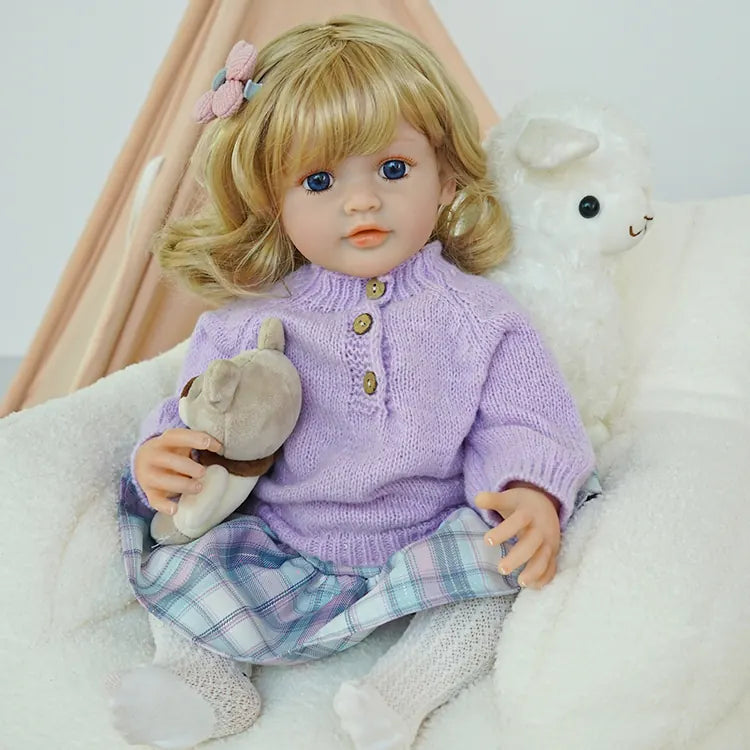 Close-up of a blonde hair, blue-eyed reborn doll wearing a purple sweater, holding a teddy bear, with a tent and sheep plush toy in the background.