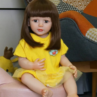 A realistic toddler doll with straight brown hair, wearing a bright yellow dress, sitting on a pink surface with a neutral expression.