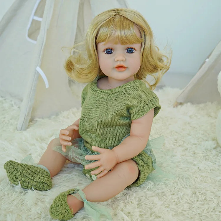 Reborn doll in green dress, holding a teddy bear, sitting on a carpet.