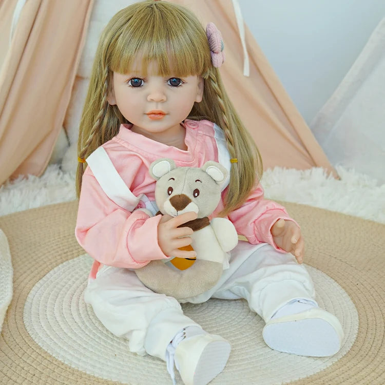 Reborn baby doll in pink shirt and white overalls, sitting on a rug, facing right.