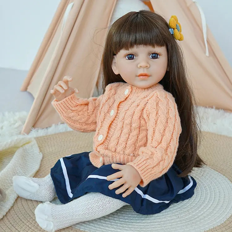 Reborn baby doll in peach sweater and navy skirt, sitting on a rug.