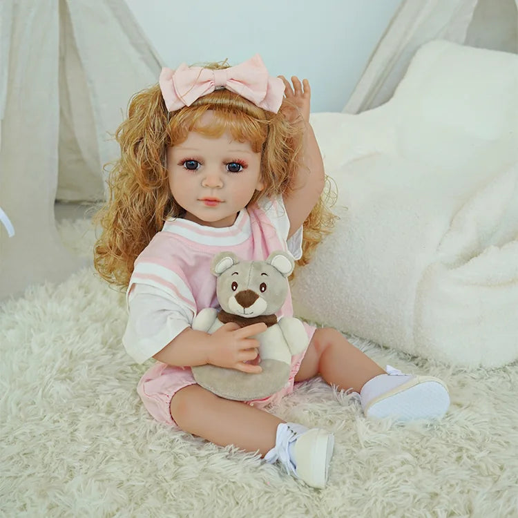 Beautiful toddler reborn doll with curly blonde hair, wearing a pink dress and bow, seated on a white fluffy chair.