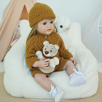 Reborn baby doll in a brown knit outfit, sitting on a rug with a teddy bear.