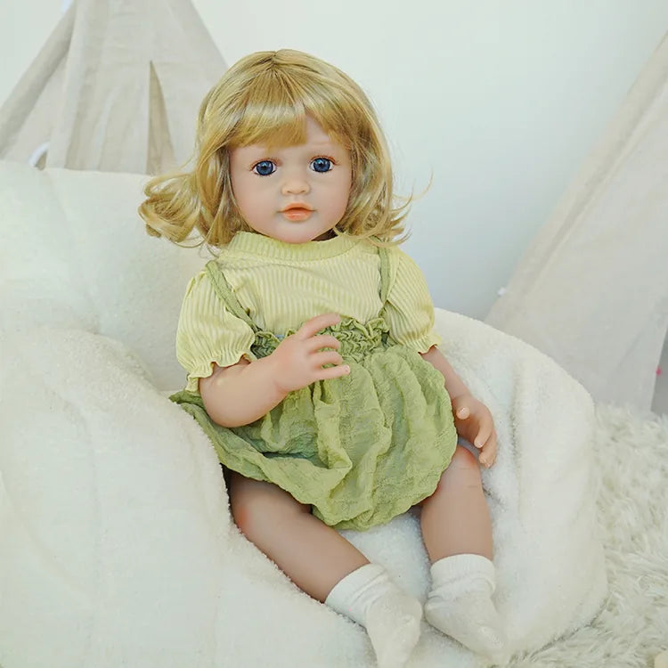 The reborn baby doll is sitting on a soft chair, showing her full-body pose with a simple indoor background.