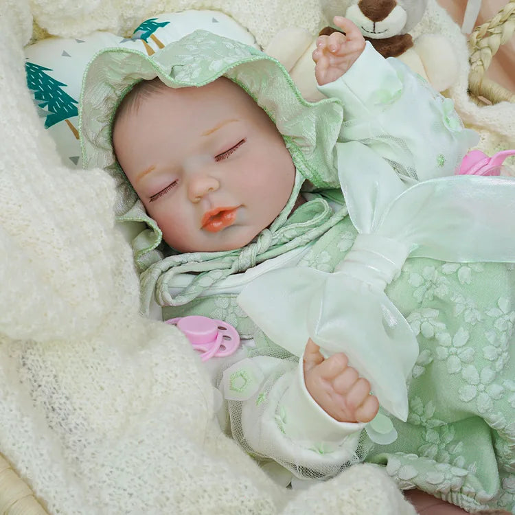 A reborn baby doll in a pale green floral hooded outfit, with a pink pacifier, sleeping snugly in a wicker basket padded with a soft white blanket. The doll is accompanied by a plush grey teddy bear and a pink bottle.
