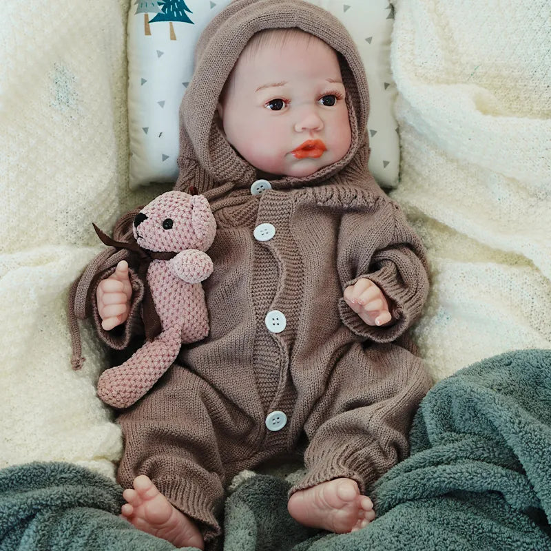 A reborn baby doll sitting up, dressed in a brown knit onesie with a hood, holding a knitted teddy bear, looking adorably lifelike.