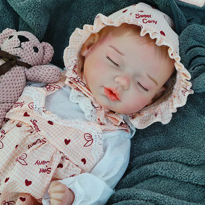 A reborn baby doll dressed in a patterned dress, holding a pink teddy bear, resting on a dark blanket.