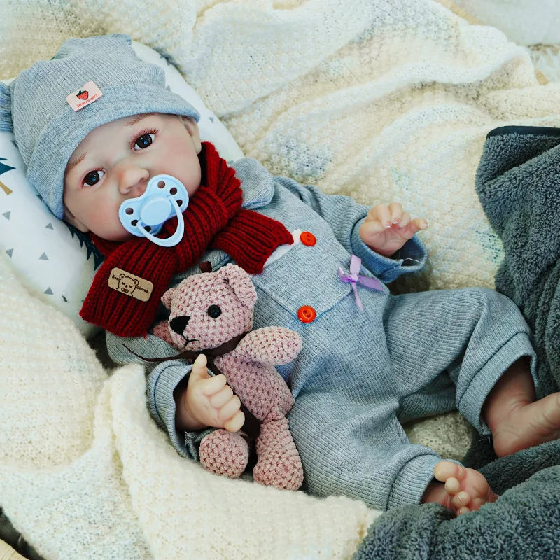 Reborn baby doll sitting up with a pacifier, dressed in a cozy gray outfit with a matching hat, holding a knitted teddy bear.