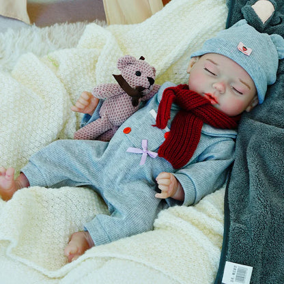 A reborn baby doll dressed in a gray outfit with a red scarf and a matching hat, lying on a light blanket, holding a pink teddy bear.