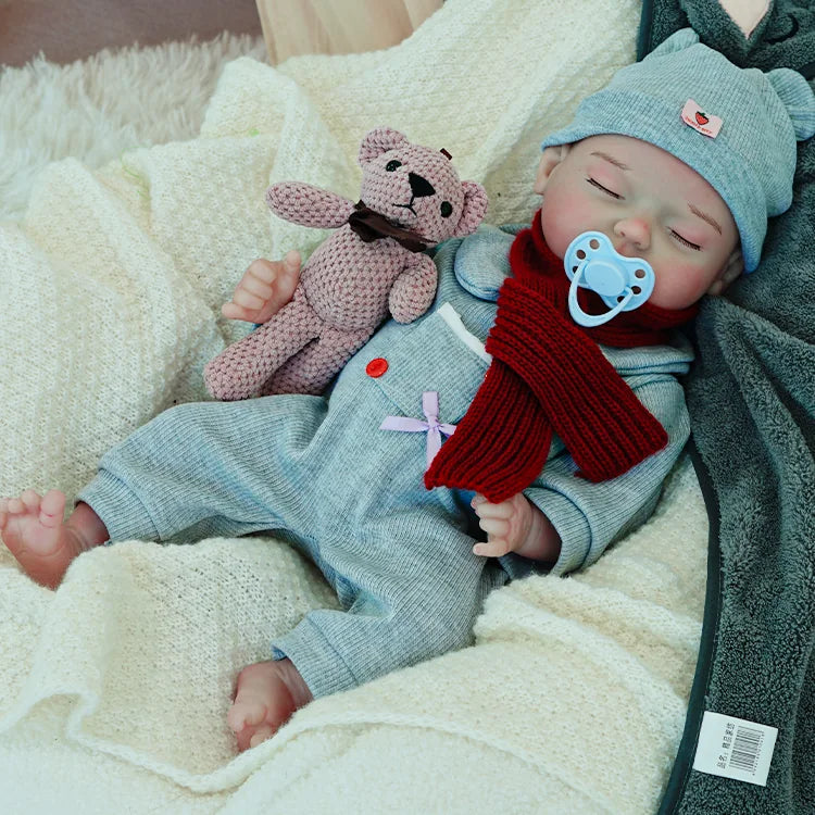 A reborn baby doll with a pacifier, dressed in a gray outfit and hat, lying on a light blanket, holding a pink teddy bear.