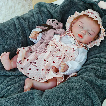 A reborn baby doll lying down, eyes closed, holding a pink teddy bear, with a dark blanket underneath.