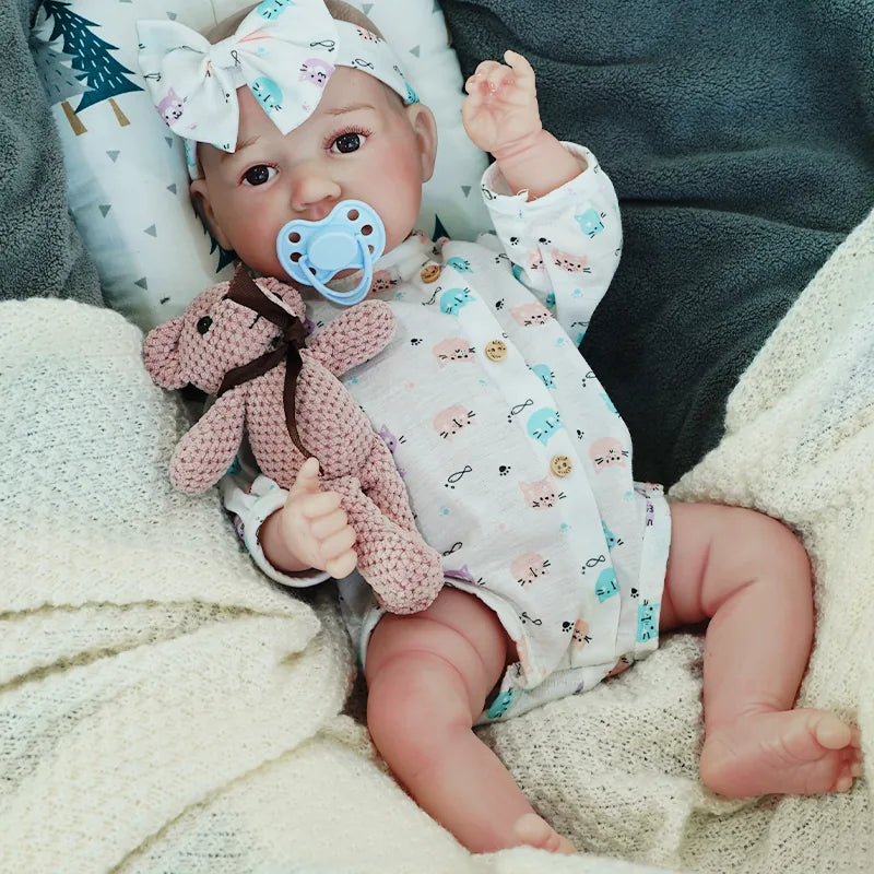 The reborn baby doll sitting up with a pacifier and a knitted teddy bear.