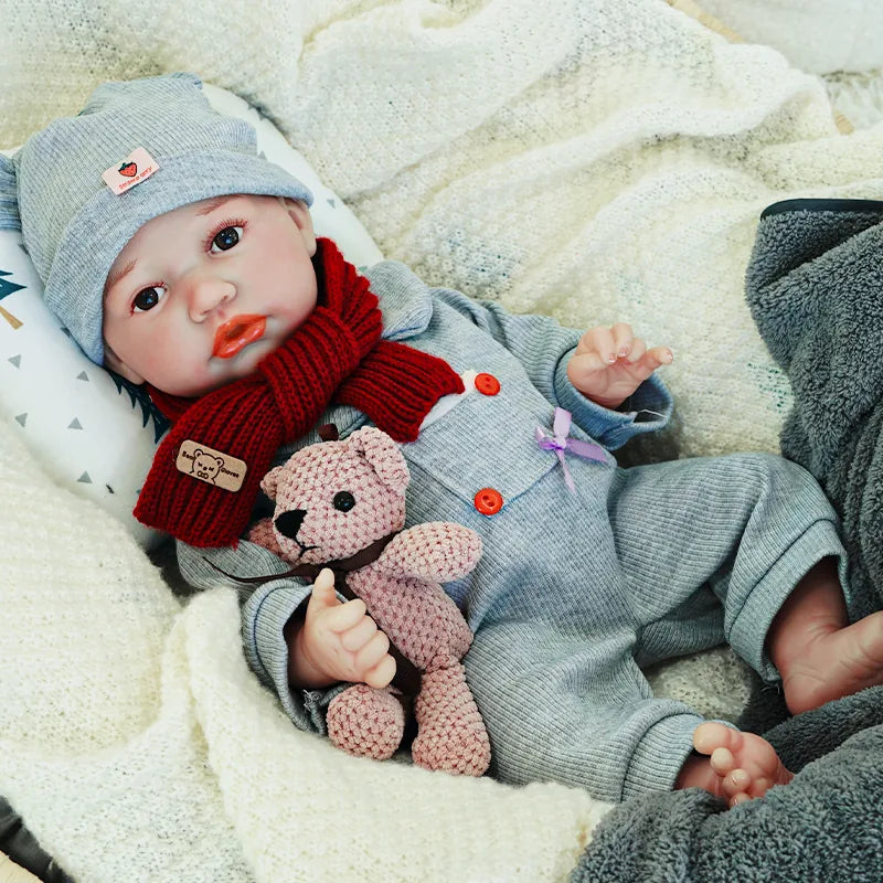 The reborn baby doll lying down in a gray onesie, red scarf, and hat, cuddling a knitted teddy bear.