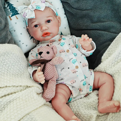 The reborn baby doll lying on a soft surface, dressed in a cute outfit and holding a teddy bear.