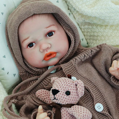 A close-up of the reborn baby doll’s face, with a serene expression, dressed in a brown hooded onesie, holding a knitted teddy bear.