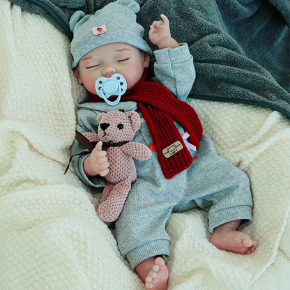 A reborn baby doll in a gray outfit with a red scarf and hat, lying on a light blanket, holding a teddy bear, arms raised.