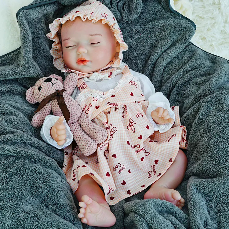 A reborn baby doll peacefully sleeping on a dark blanket, holding a pink teddy bear.