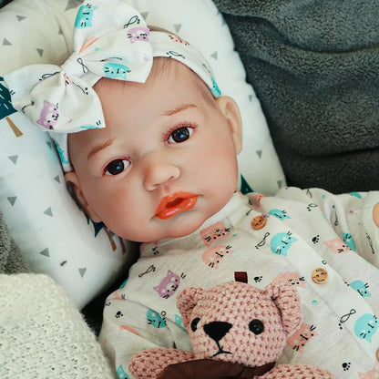 Reborn baby doll lying down in a cute onesie with a bow headband, holding a knitted teddy bear.